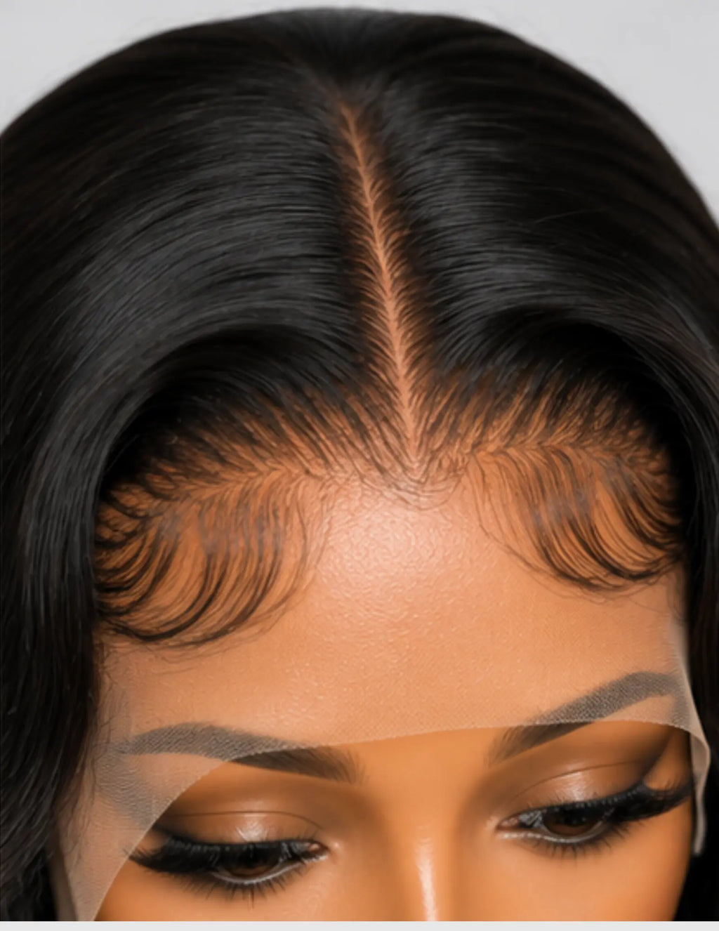 Close-up of a person wearing a black lace front wig, showing the hair part, hairline, and eyelashes against a light gray background.